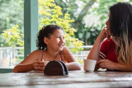 kleine brünette latina Mädchen lächelt, während sie eine Tasse Kaffee neben ihrer Mutter, Mutter und Tochter im Gespräch trinkt