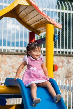 Photo for Happy little latina brunette girl smiling about to go down the slippery slide in the playground. playground equipment - Royalty Free Image