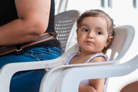 petite fille latina, assise sur une chaise blanche ennuyée regardant vers le côté tout en buvant de l'eau d'un verre