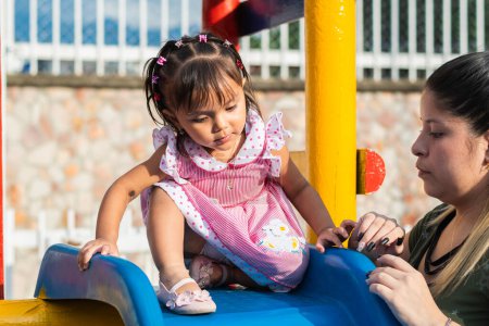 Photo for Little brunette latina girl with her mother about to go down the small slippery slide in the park. - Royalty Free Image