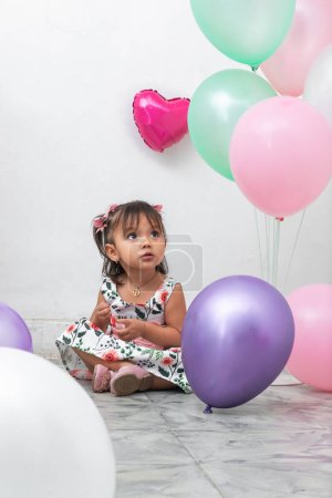 little brunette latina girl sitting on the ground surrounded by colorful balloons, looking upwards