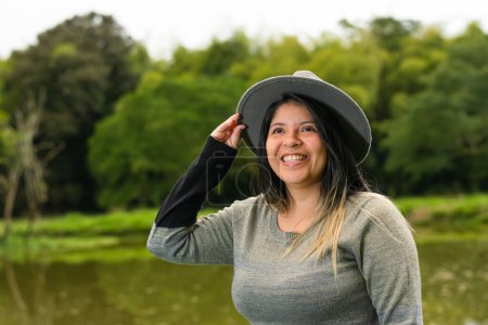 Téléchargez les photos : Belle latina femme souriant et levant les yeux tout en tenant son chapeau et posant près d'un lac - en image libre de droit
