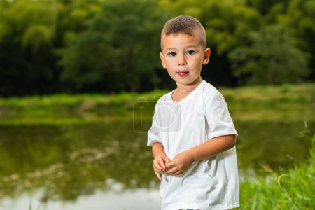 pequeño chico latino mirando curiosamente mientras está de pie con un lago de pesca en el fondo