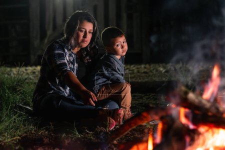 Photo for Young latin woman carrying her son, sitting in front of a campfire at night, looking sad and bored. - Royalty Free Image