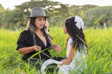 joven madre latina con su hija pequeña, sentada en la hierba alta hablando de la vida