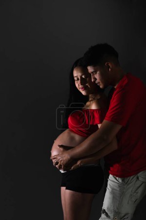 junges lateinisches Paar, Mann umarmt seine Frau von hinten, während er ihren Bauch berührt und glücklich auf ihr Baby wartet