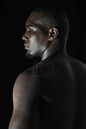 Nahaufnahme des Rückens eines schwarzen afroamerikanischen Mannes, der aus dem Augenwinkel auf einen schwarzen Hintergrund mit 90-Grad-Beleuchtung blickt.