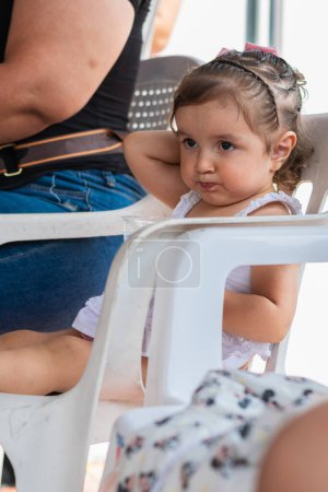 little latina girl, sitting on a white chair scratching her head while waiting for food