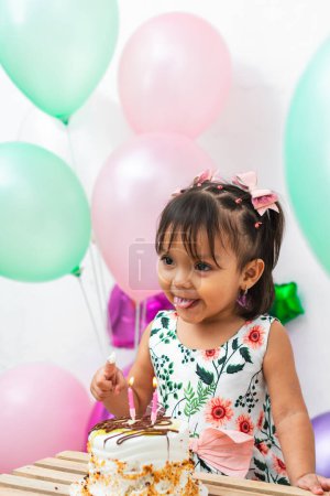 petite fille brune latine étalant son doigt avec du gâteau pour l'essayer, entourée de ballons colorés
