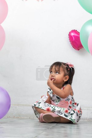 little brunette latina girl sitting on the ground surrounded by balloons, putting a hand to her mouth while looking up