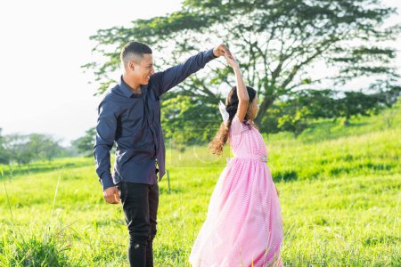 latin father holding his daughter's hand, while she dances, standing on a grass field, smiling.