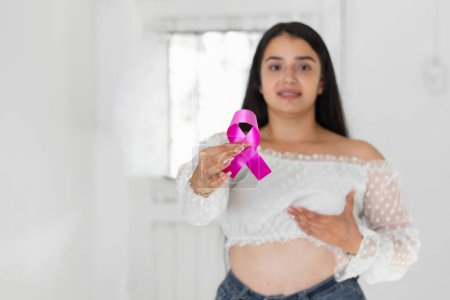 caucasian latina woman, showing a pink ribbon, symbol of breast cancer, while touching her breast with her other hand.