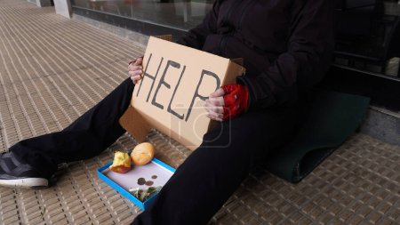 Photo for A homeless beggar woman asks for alms sitting at the entrance to a cafe with a sign: HELP. At her feet is a box into which people throw coins and food - Royalty Free Image