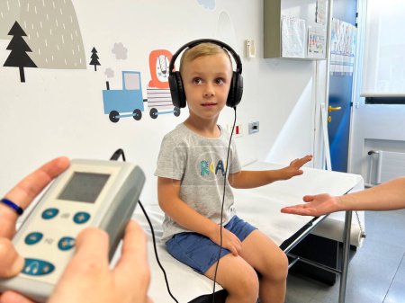 un niño rubio pequeño sentado con auriculares sometidos a una prueba de audición en una clínica