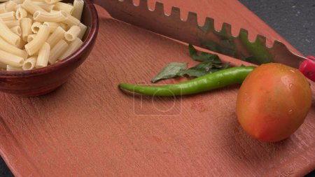 Photo for Fresh raw pasta in wooden bowl with green chili, knife and tomato on brown cutting board - Royalty Free Image