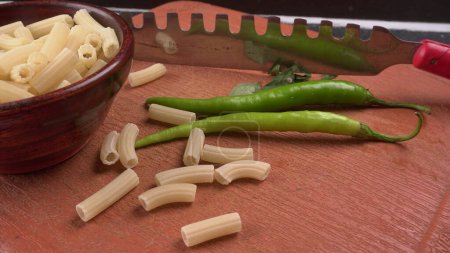 Photo for Fresh raw pasta in wooden bowl with green chili, knife and tomato on brown cutting board - Royalty Free Image
