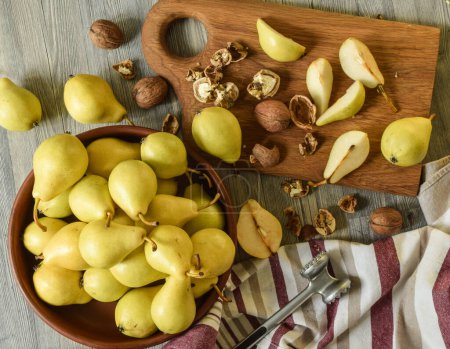 Photo for Top table view of ripe yellowish pears in clay bowl on wooden table indoors. Sliced pear pieces, split walnuts on wooden cutting board. Kitchen mallet hammer lying on traditional dishtowel. Still life - Royalty Free Image