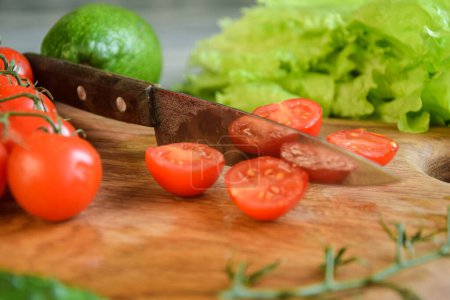 Photo for Closeup kitchen knife, sliced tomatoes on wooden cutting board on background of green vegetables indoors. Food cooking background. Ingredients for preparation salad dishes. Healthy food concept - Royalty Free Image