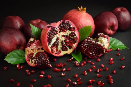 Photo for Split open fruit scattered red grains seed indoors. Still life ripe pomegranates, red apples, green leaves on black background. Cluster with fresh pomegranate arils. Studio shot. Health nutrition - Royalty Free Image