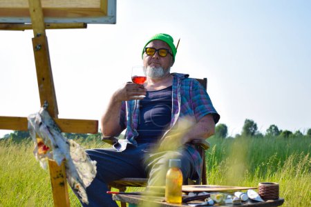 Photo for Offbeat mature artist relaxing after completion artwork at field, drinks wine looking at picture sitting on chair outdoors in morning sunshine. Canvas on wooden easel painting tools materials supplies - Royalty Free Image