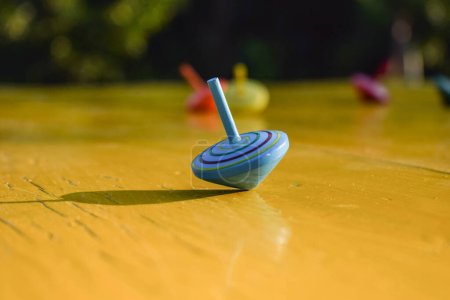 Foto de Close-up solo spinning top color azul gira en la mesa amarilla, mientras que otros juguetes superiores cayeron al aire libre. Objeto giratorio como analogía para equilibrar la vida. hermoso paisaje parque dorado sol rayos fondo - Imagen libre de derechos