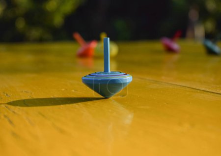 Foto de Close-up solo spinning top color azul gira en la mesa amarilla, mientras que otros juguetes superiores cayeron al aire libre. Objeto giratorio como analogía para equilibrar la vida. hermoso paisaje parque dorado sol rayos fondo - Imagen libre de derechos