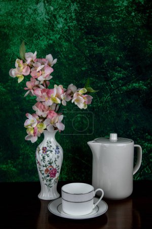Photo for Artificial flowers in a white decorated porcelain vase with white teapot and teacup and saucer on a wooden table top with mottled background - Royalty Free Image
