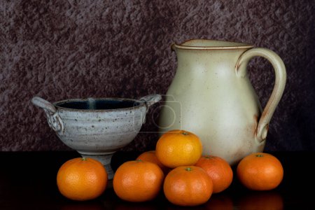 Foto de Jarra de vino de cerámica y tazón de cáliz con naranjas satsuma en una superficie de madera pulida - Imagen libre de derechos