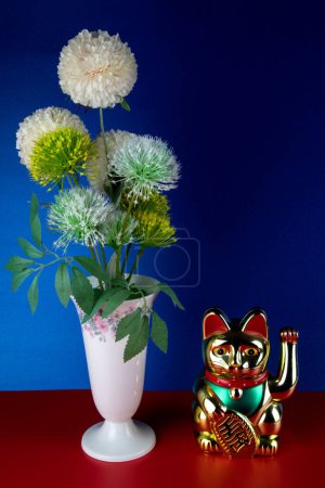 Porzellanvase mit künstlichen Blumen und goldfarbener Maneki neko winkt Glückskatze