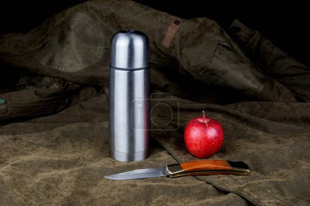 Vacuum flask with open knife and red apple on an outdoor field coat