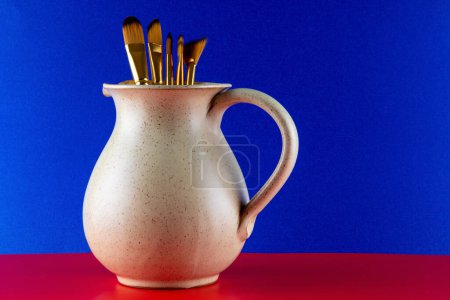 Artists paint brushes in an earthenware wine jug on a blue background and red base