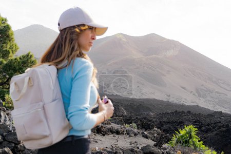 Photo for Pensive backpacker woman with cap looks towards the Tajogaite volcano, sideways, she looks out of focus in the foreground, from a viewpoint on the island of La Palma - Royalty Free Image