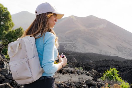 Photo for Smiling backpacker woman with cap looks towards the Tajogaite volcano, sideways, the volcano is seen out of focus in the background, from a viewpoint on the island of La Palma - Royalty Free Image