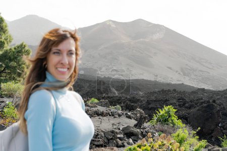 Photo for Backpacker woman smiles looking aside at the Tajogaite volcano, she is seen out of focus in the foreground, on the island of La Palma - Royalty Free Image