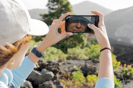 Photo for Close-up woman with a cap taking a photo of the Tajogaite volcano with her cell phone and she sees herself reflected in the phone - Royalty Free Image