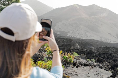 Photo for Woman with a cap taking a vertical photo of the Tajogaite volcano with her cell phone and she sees herself reflected in the phone, she comes out of focus in the foreground - Royalty Free Image