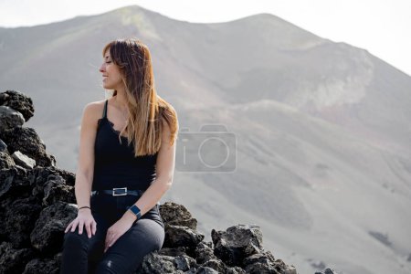 Photo for Woman in suspenders looks at the ground smiling in front of the Tajogaite volcano, the volcano is seen out of focus in the background, at a viewpoint on the island of La Palma - Royalty Free Image