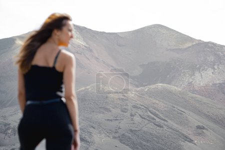 Photo for Close-up woman standing with hair blowing in the wind sideways in front of the Tajogaite volcano from a viewpoint, on top of some rocks, on the island of La Palma - Royalty Free Image