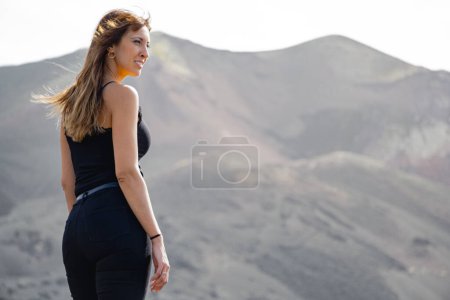 Photo for Pensive woman standing with her hair blowing in the wind sideways in front of the Tajogaite volcano from a viewpoint, on top of some rocks, on the island of La Palma - Royalty Free Image
