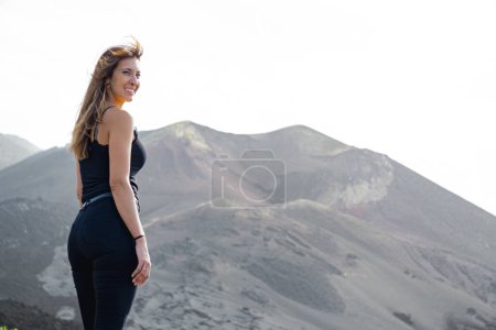 Photo for Standing woman smiles from the viewpoint of the Tajogaite volcano, on the island of La Palma, the volcano is seen out of focus in the background - Royalty Free Image