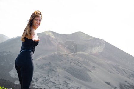 Photo for Standing woman smiles, extends her hand looking at the camera, with her hair flying in the wind, in front of the Tajogaite volcano, from a viewpoint, on the island of La Palma - Royalty Free Image