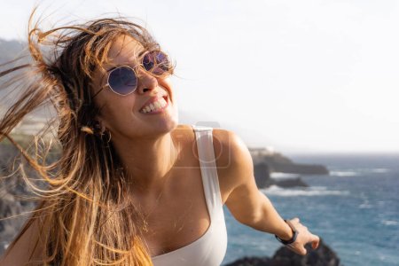 woman with sunglasses smiling looking at the camera, with her hair blown by the wind, at a viewpoint on a beach in La Palma