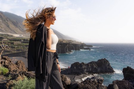 woman poses with sunglasses, a jacket and hair blown by the wind on some cliffs in front of a beach in La Palma