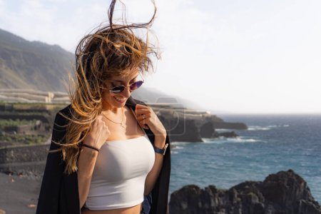 woman smiling puts on a jacket with hair blown by the wind on some cliffs in front of a beach in La Palma, looking down