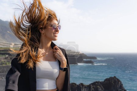 smiling woman puts on a jacket with hair blown by the wind on some cliffs in front of a beach in La Palma, sideways