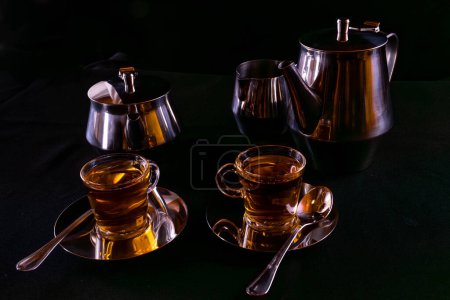 Two glass cups filled with tea on silver-plated metal plates with containers to store the liquid