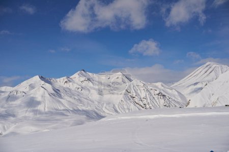Foto de Aérea de pistas de montaña nevadas llenas de polvo para freeride en la estación de esquí en el amanecer de invierno. Cordillera de campiña cubierta de nieve virgen. Picos del Cáucaso horizonte con un resplandor de crepúsculo. - Imagen libre de derechos