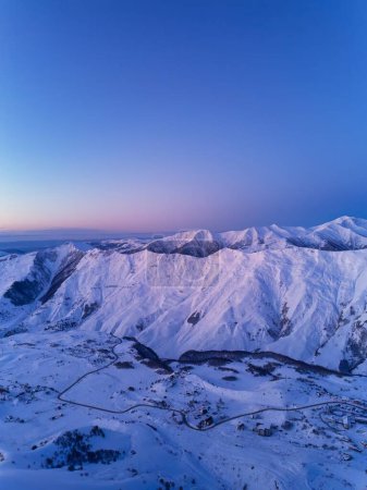 Photo for Aerial of snowy mountain range on winter sunrise at ski resort. Moon above mountains valley and village with switchbacks road at a sunset. Caucasus peaks skyline in the dusk. City lights at night. - Royalty Free Image