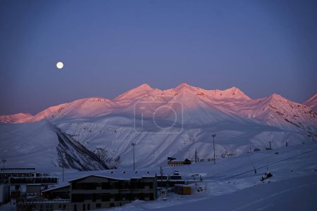 Photo for Aerial of snowy mountain range on winter sunrise at ski resort. Moon above mountains valley and village with switchbacks road at a sunset. Caucasus peaks skyline in the dusk. City lights at night. - Royalty Free Image