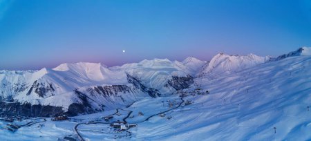 Photo for Wide aerial panorama of snowy mountain ridge on winter sunrise. Stunning mountains range covered with snow powder on ski resort at sunset. Moon above caucasus peaks skyline in a dusk at night. Gudauri - Royalty Free Image